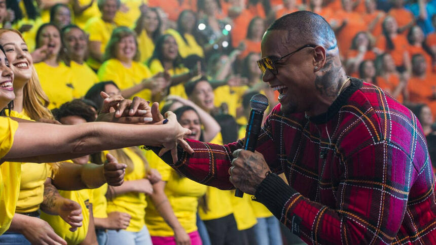 Leo Santana de camisa quadriculada em tons de preto e magenta, sorrindo, segurando a mão de uma fã na plateia do Faustão na Band