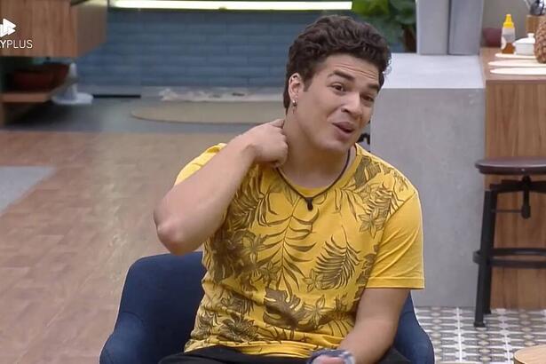 Lucas sentando na poltrona com uma camisa estampada e coçando o pescoço.