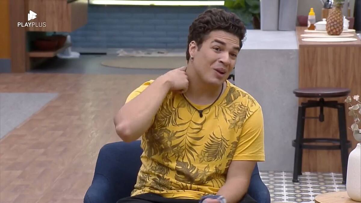 Lucas sentando na poltrona com uma camisa estampada e coçando o pescoço.