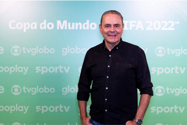 Luís Roberto com uma roupa preta, em frente a um banner verde de divulgação da Copa na Globo