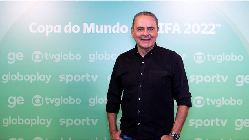 Luís Roberto com uma roupa preta, em frente a um banner verde de divulgação da Copa na Globo