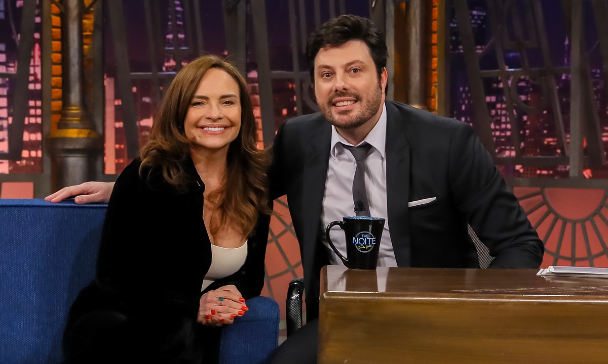 Luiza Tomé de blazer e calça preta, camisa branca por dentro, sorrindo, sentada num sofá azul ao lado de Danilo Gentili durante gravação do The Noite, do SBT