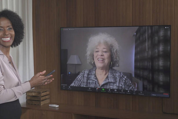 Maju Coutinho, com uma calça preta e uma blusa com tom mais claro, de sorriso aberto e um celular na mão. Ela está em um estúdio, gravando uma entrevista com Angela Davis, através de uma televisão