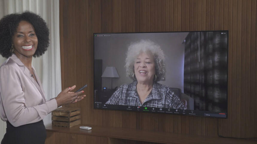 Maju Coutinho, com uma calça preta e uma blusa com tom mais claro, de sorriso aberto e um celular na mão. Ela está em um estúdio, gravando uma entrevista com Angela Davis, através de uma televisão