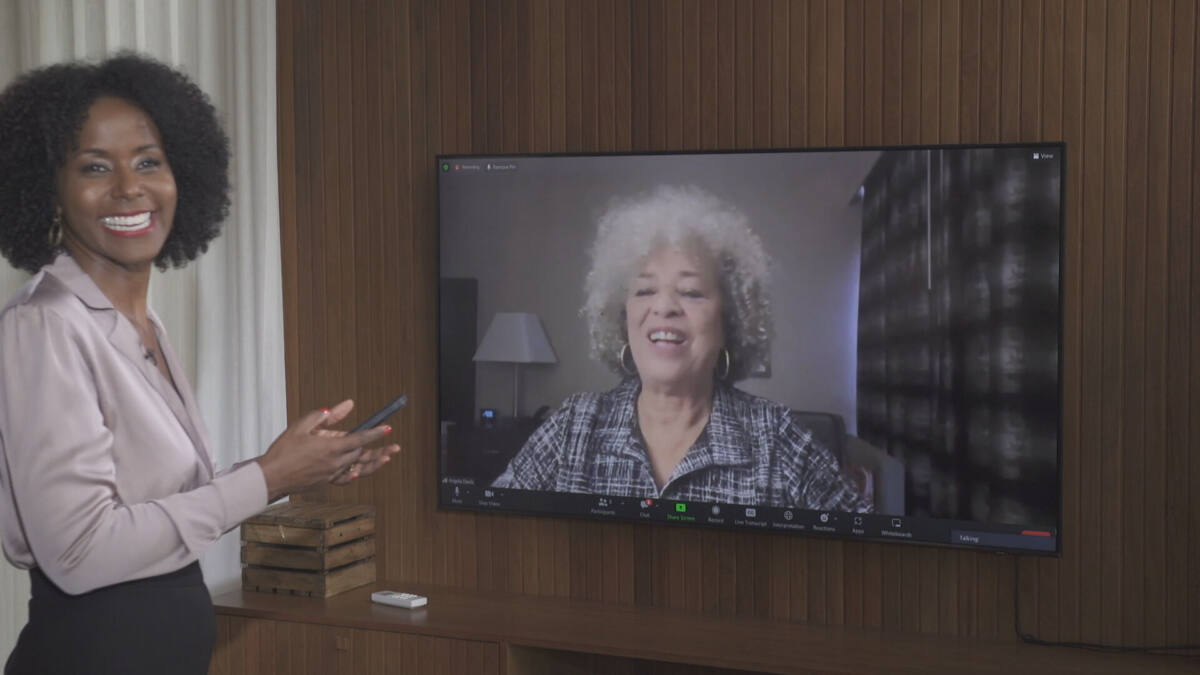 Maju Coutinho, com uma calça preta e uma blusa com tom mais claro, de sorriso aberto e um celular na mão. Ela está em um estúdio, gravando uma entrevista com Angela Davis, através de uma televisão