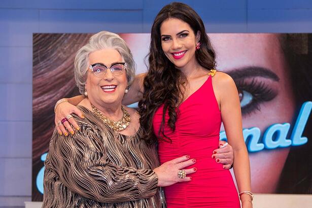 Mamma Buschetta de vestimo marrom, sorrindo, com as mãos na cintura de Daniela Albuquerque, que está de vestido e batom vermelho, sorrindo