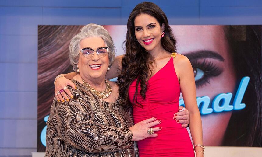 Mamma Buschetta de vestimo marrom, sorrindo, com as mãos na cintura de Daniela Albuquerque, que está de vestido e batom vermelho, sorrindo