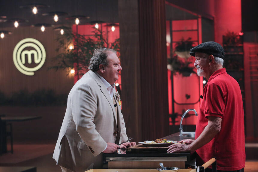 Jacquin, com um terno cinza, em frente a bancada do MasterChef+ com o participante Astro, utilizando uma roupa vermelha e uma boina na cabeça