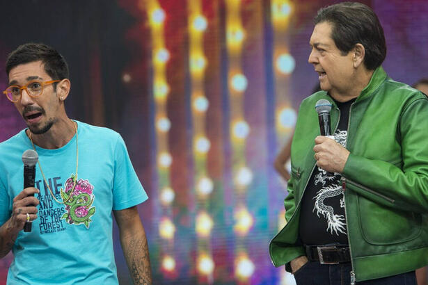 Mateus Cidrão de camisa azul, segurando o microfone, sorrindo, ai lado de Faustão durante gravação de programa