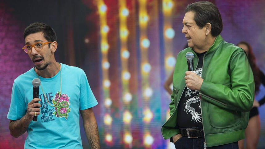 Mateus Cidrão de camisa azul, segurando o microfone, sorrindo, ai lado de Faustão durante gravação de programa