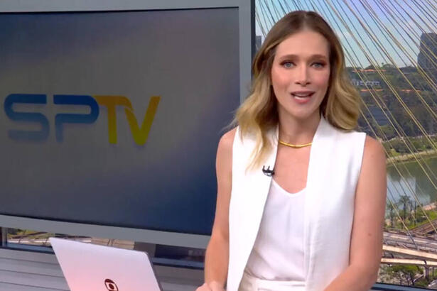 Natália Ariede de roupa branca, do estúdio do SP1, em frente a uma televisão e apoiada em um púlpito com um notebook