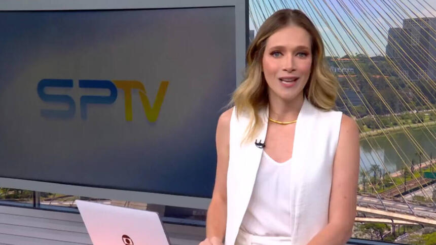 Natália Ariede de roupa branca, do estúdio do SP1, em frente a uma televisão e apoiada em um púlpito com um notebook