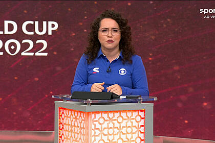 Natália Lara, com um uniforme de esporte da Globo, em frente ao púlpito, com um telão ao fundo