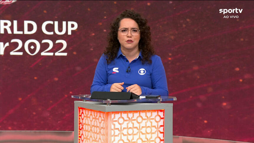 Natália Lara, com um uniforme de esporte da Globo, em frente ao púlpito, com um telão ao fundo