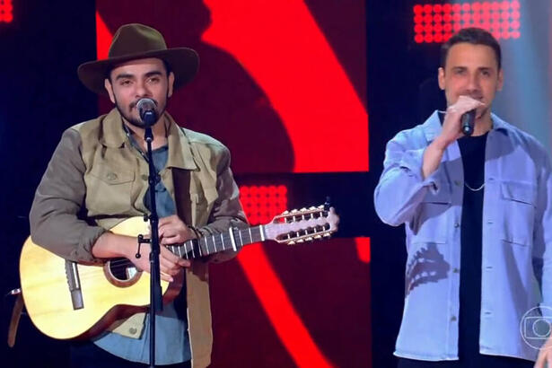 Dupla Neto & Felipe cantando no palco do The Voice Brasil. O primeiro toca um violão e tem um chapéu na cabeça. O segundo segura um microfone de mão