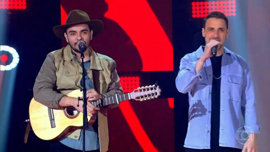 Dupla Neto & Felipe cantando no palco do The Voice Brasil. O primeiro toca um violão e tem um chapéu na cabeça. O segundo segura um microfone de mão