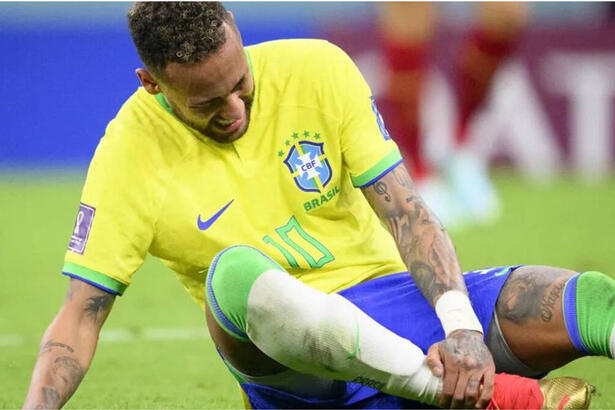 Neymar, sentado no gravado, com a mão no tornozelo, com cara de sofrimento e chorando. Ele usa com camisa da Seleção Brasileira