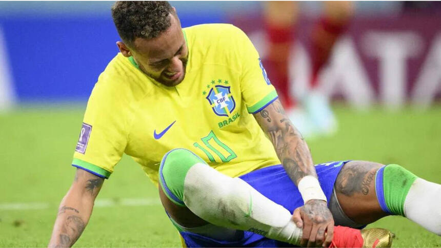 Neymar, sentado no gravado, com a mão no tornozelo, com cara de sofrimento e chorando. Ele usa com camisa da Seleção Brasileira