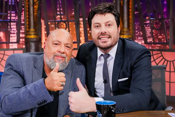 Nilson César de blazer cinza e calça preta, camisa branca por dentro, sorrindo, sentada num sofá azul ao lado de Danilo Gentili durante gravação do The Noite, do SBT