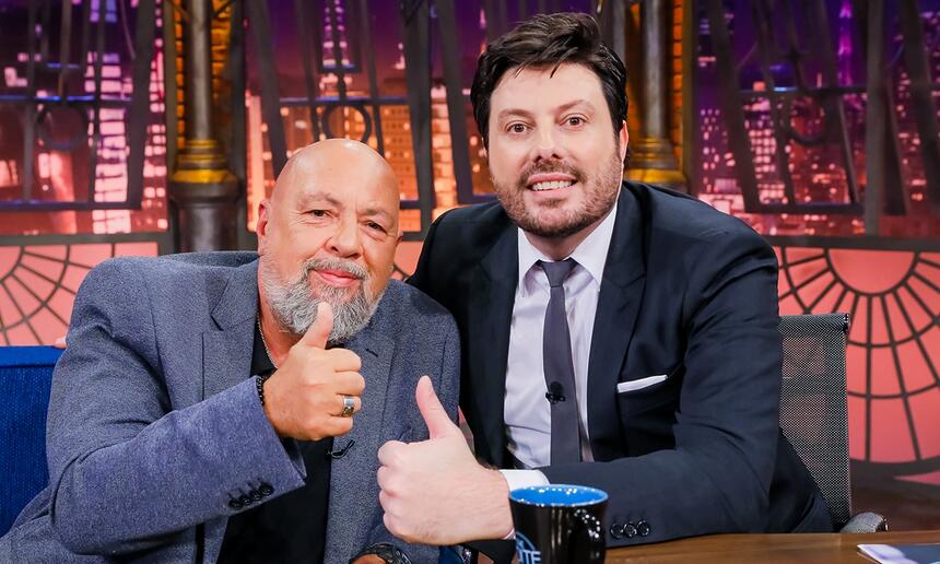 Nilson César de blazer cinza e calça preta, camisa branca por dentro, sorrindo, sentada num sofá azul ao lado de Danilo Gentili durante gravação do The Noite, do SBT