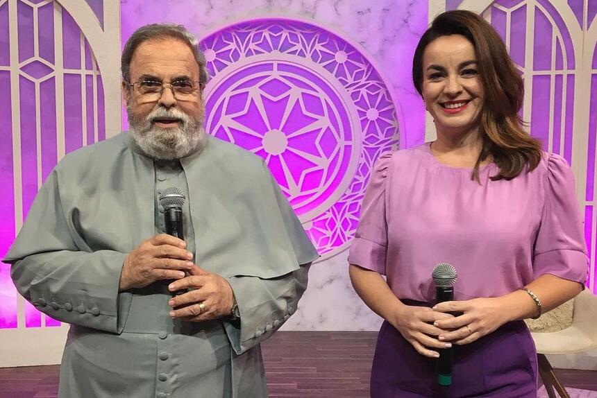 Pe Antonio Maria de bata cinza segurando um microfone e Dani Espíndola de camisa roxa e calça preta sorrindo durante gravação do Sábado com Maria, da TV Aparecida