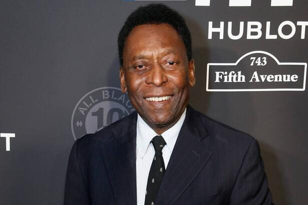 Pelé com um terno e gravata, sorrindo, em foto posada em frente e um banner preto de um evento