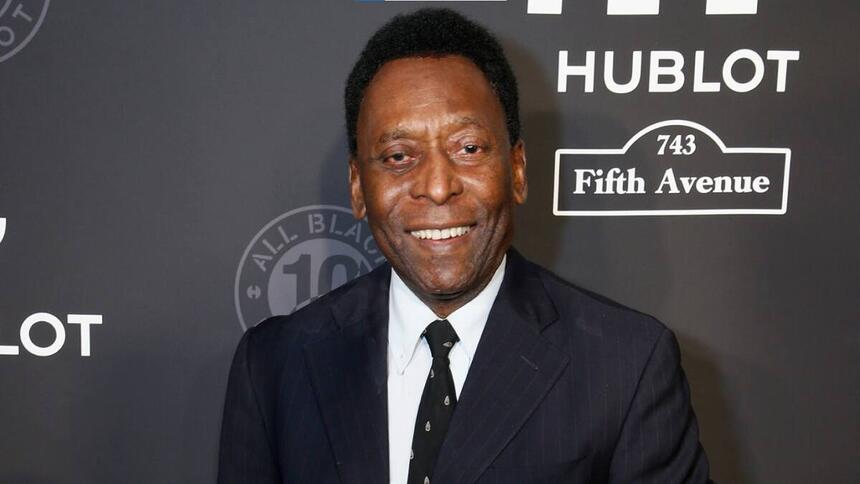 Pelé com um terno e gravata, sorrindo, em foto posada em frente e um banner preto de um evento