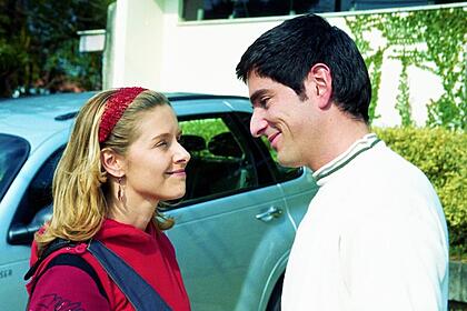 Julia (Bianca Rinaldi) sorrir para Alberto (Rodrigo Veronese) em um estacionamento, próximo de um carro