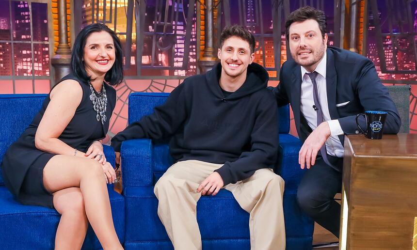 Pkllipe e Tuca de camisa preta, sorrindo, sentados num sofá azul ao lado de Danilo Gentili para gravação do The Noite, do SBT