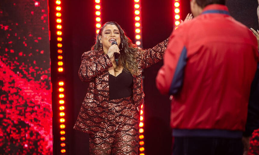 Preta Gil de blazer a calça vermelha com lantejolas, camisa preta, com os braços abertos segurando o microfone com a mão direita durante gravação do Faustão na Band