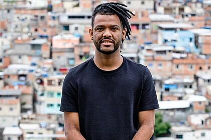 Rene Silva de camisa básica preta, sorrindo, de cabelo trançado preso sentado durante ensaio fotográfico no Morro