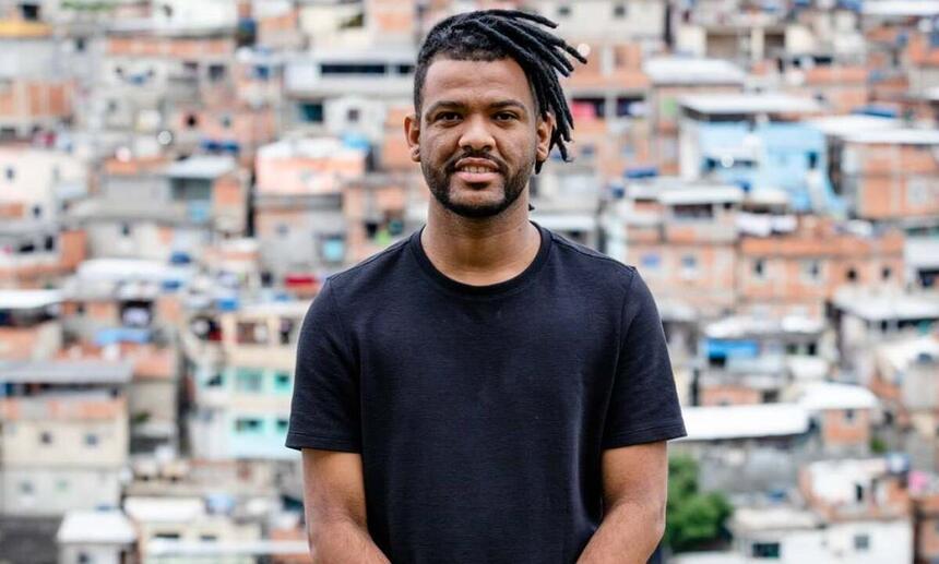 Rene Silva de camisa básica preta, sorrindo, de cabelo trançado preso sentado durante ensaio fotográfico no Morro