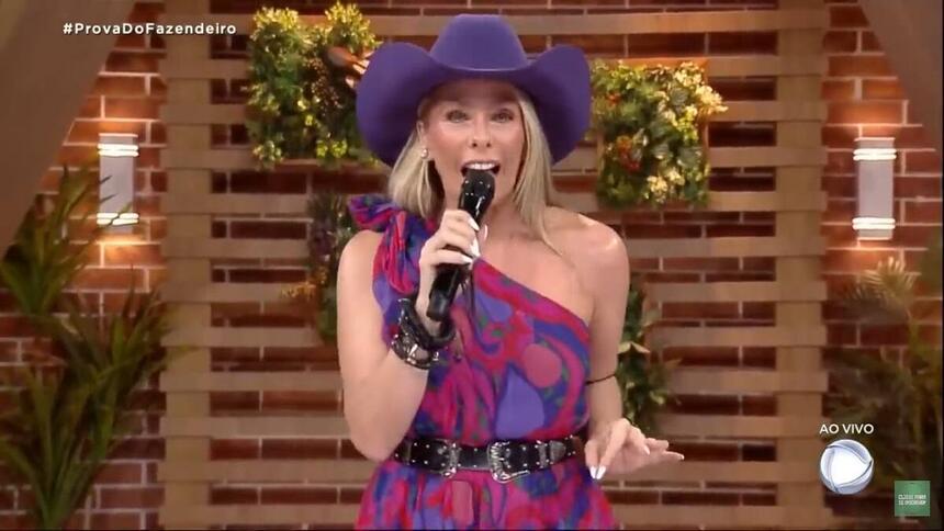 Adriane com o microfone na mão, com uma roupa estampada e um chapéu de cor lilás.