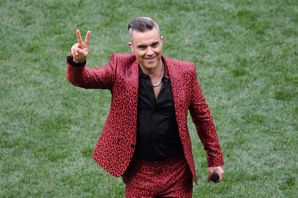 Robbie Williams de paletó e calça vermelhos, camisa preta fazendo um 2 com a mão direita e segurando um microfone preto durante performance nas Olimpiadas