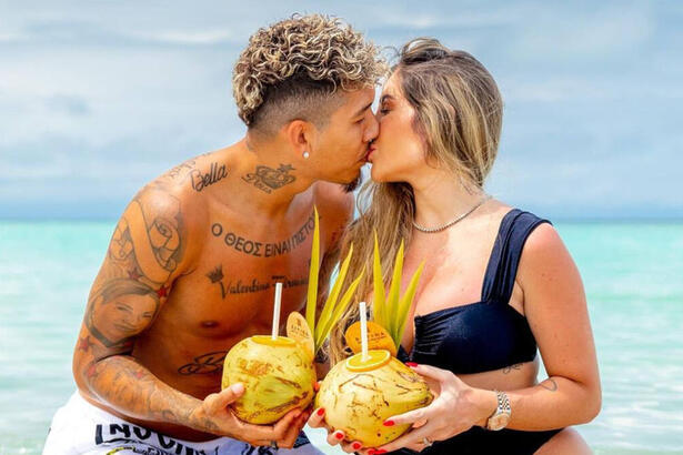 Roberto Firmino, sem camisa, beijando a esposa. Ambos seguram um coco nas mãos, com uma praia ao fundo