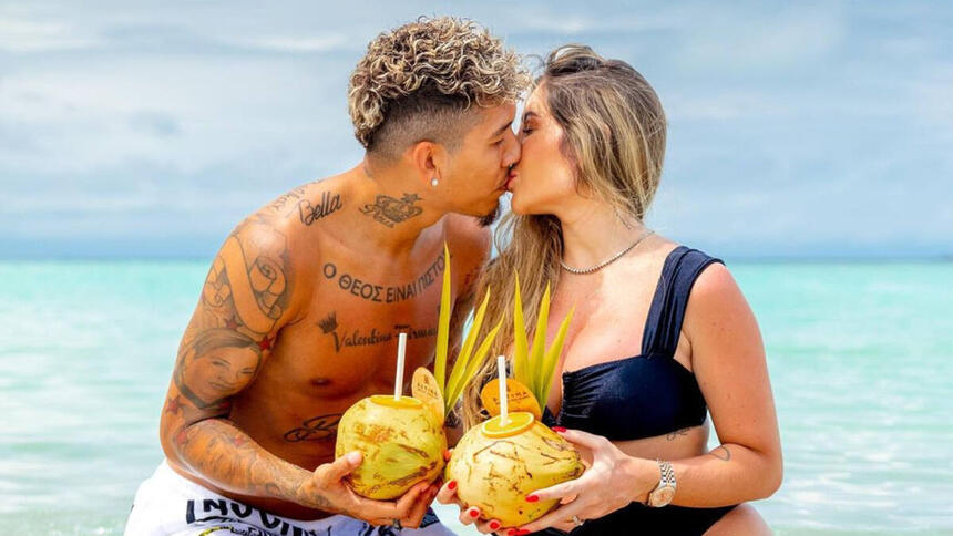 Roberto Firmino, sem camisa, beijando a esposa. Ambos seguram um coco nas mãos, com uma praia ao fundo