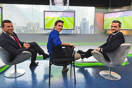 Rodrigo Bocardi ao lado dos colegas Alessandro Jodar e Tiago Scheuer, sentados no estúdio do Bom Dia Brasil, com os televisores ligados no jogo da Copa
