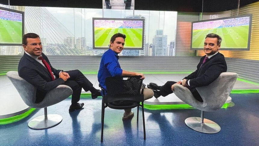 Rodrigo Bocardi ao lado dos colegas Alessandro Jodar e Tiago Scheuer, sentados no estúdio do Bom Dia Brasil, com os televisores ligados no jogo da Copa