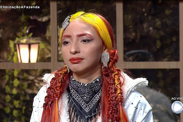 Ruivinha de Marte com camisa branca, cachecol cinza, cabelo partido ao meio, de batom vermelho, sentada na sede de eliminação de A Fazenda, da Record TV