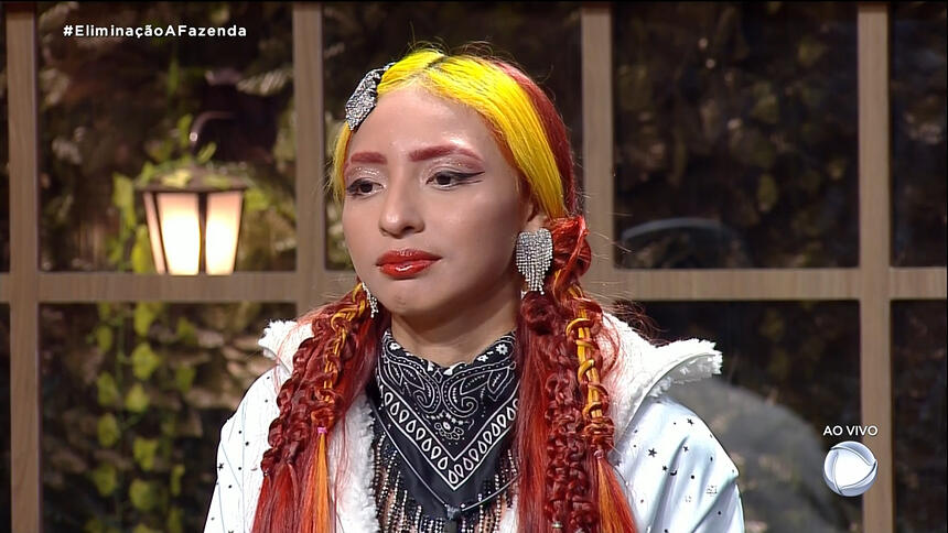 Ruivinha de Marte com camisa branca, cachecol cinza, cabelo partido ao meio, de batom vermelho, sentada na sede de eliminação de A Fazenda, da Record TV