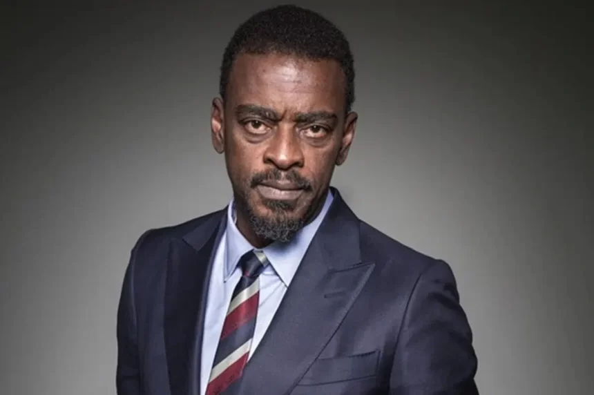 Seu Jorge com semblante sério, de paletó preto, gravata listrada para ensaio fotográfico da revista Veja