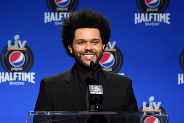 The Weeknd sorrindo, de camisa social preta e jaqueta de veludo preta em frente a um púlpito durante coletiva do Super Bowl