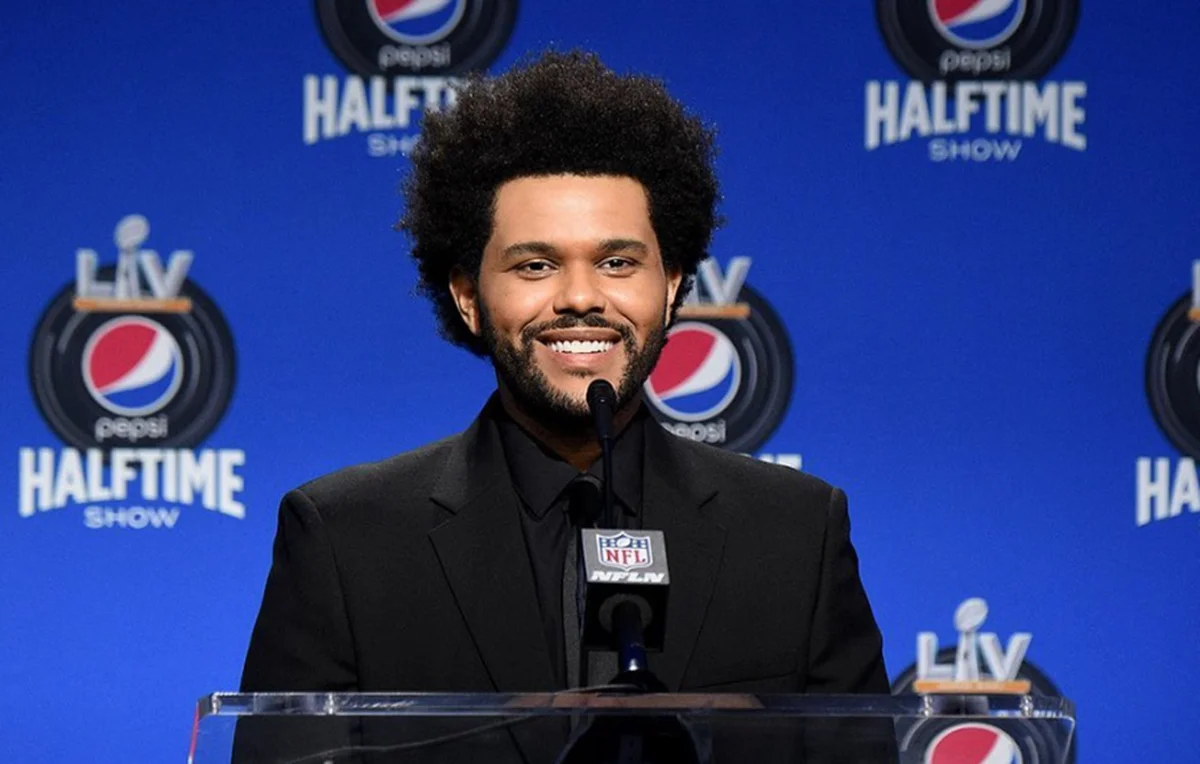 The Weeknd sorrindo, de camisa social preta e jaqueta de veludo preta em frente a um púlpito durante coletiva do Super Bowl