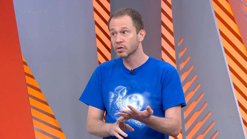 Tiago Leifert, com uma camisa azul nos estúdios do Globo Esprote, desticulando com as mãos e olhando para o seu lado direito