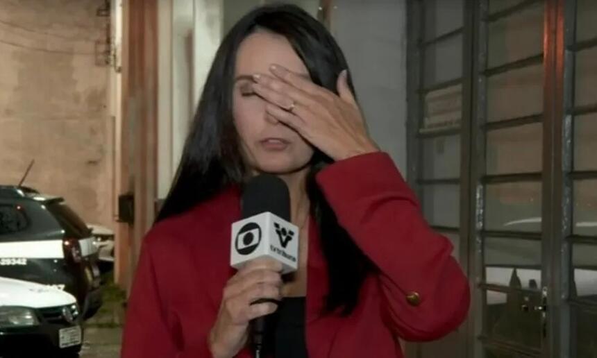 Vanessa Medeiros de cardigan vermelho e camisa preta segurando o microfone e com a mão na testa