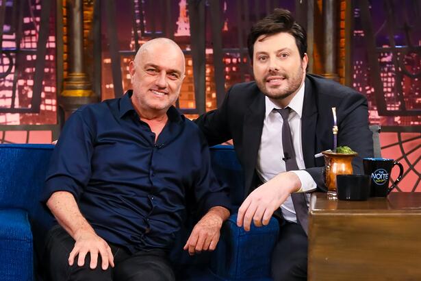 Wener Schunneman de camisa azul e calça preta, camisa branca por dentro, sorrindo, sentada num sofá azul ao lado de Danilo Gentili durante gravação do The Noite, do SBT