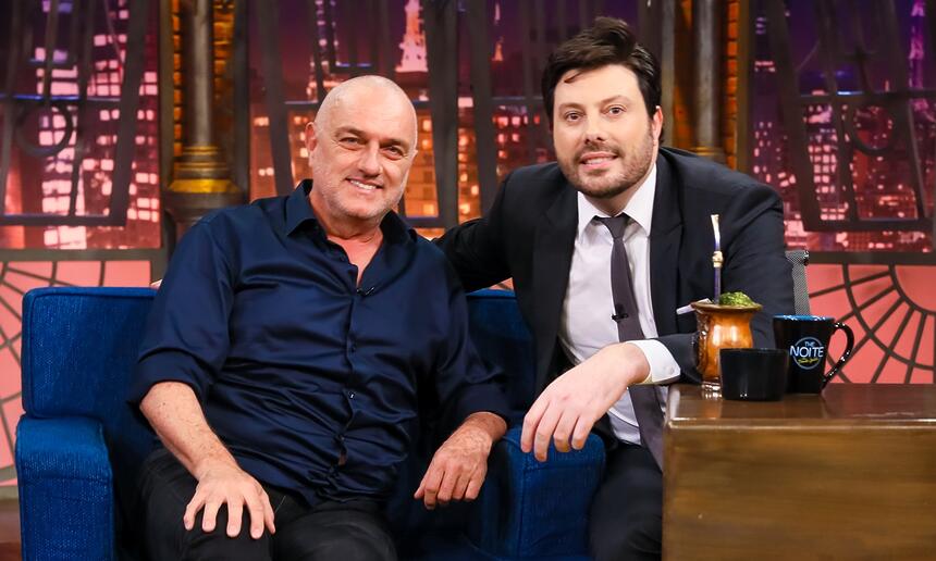 Wener Schunneman de camisa azul e calça preta, camisa branca por dentro, sorrindo, sentada num sofá azul ao lado de Danilo Gentili durante gravação do The Noite, do SBT