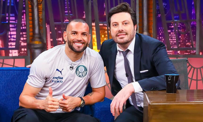 Weverton Pereira sorrindo, de camisa cinza, fazendo legal com as duas mãos, sentado ao lado de Danilo Gentili durante gravação do The Noite, do SBT