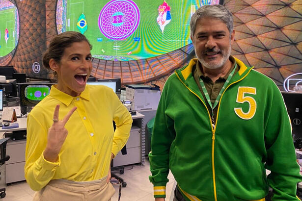 William Bonner e Renata Vasconcellos, vestidos com roupa verde e amarela, na redação do JN, com telão ao fundo com o jogo da Copa