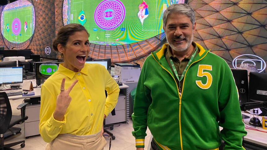 William Bonner e Renata Vasconcellos, vestidos com roupa verde e amarela, na redação do JN, com telão ao fundo com o jogo da Copa